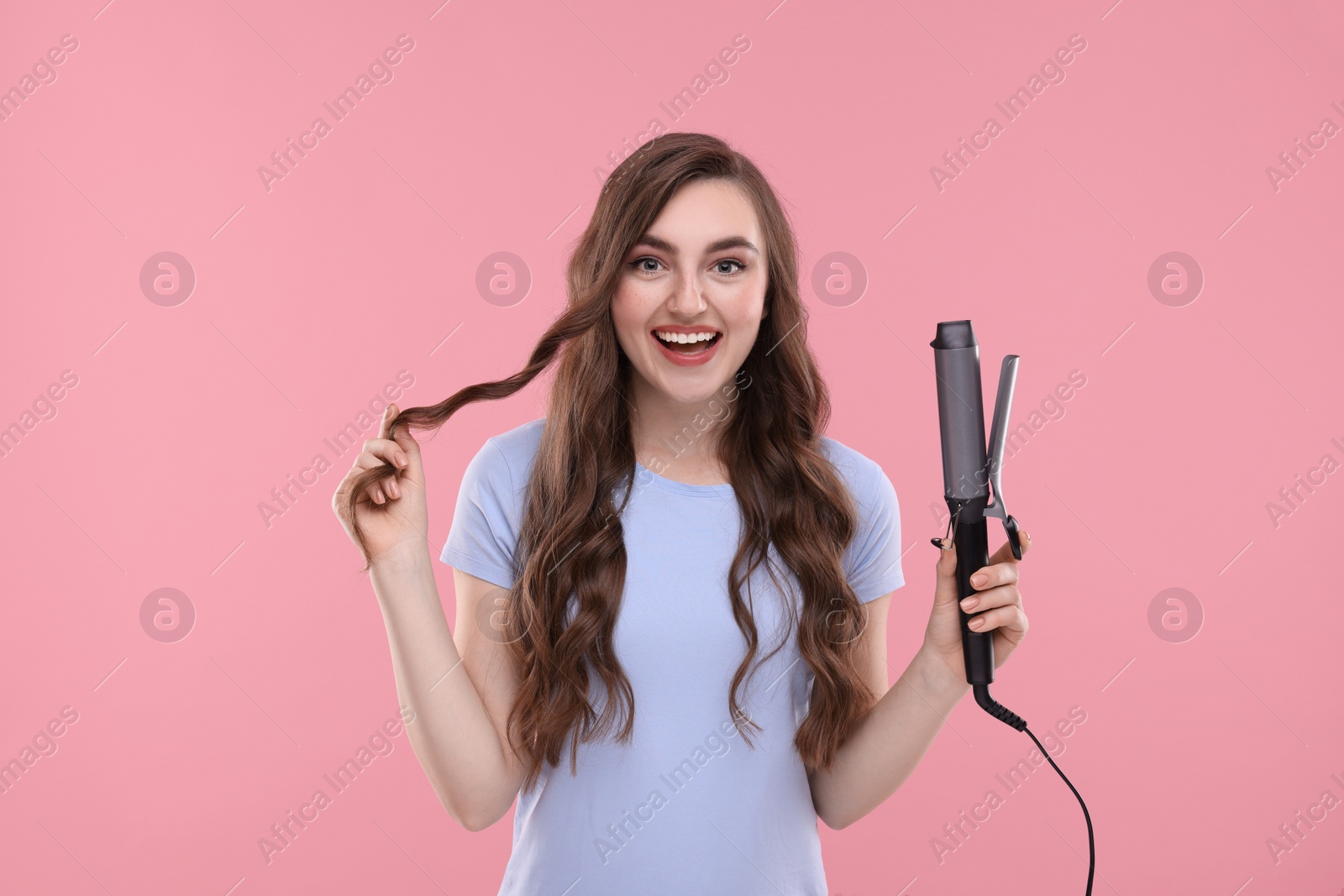 Photo of Happy young woman with beautiful hair holding curling iron on pink background