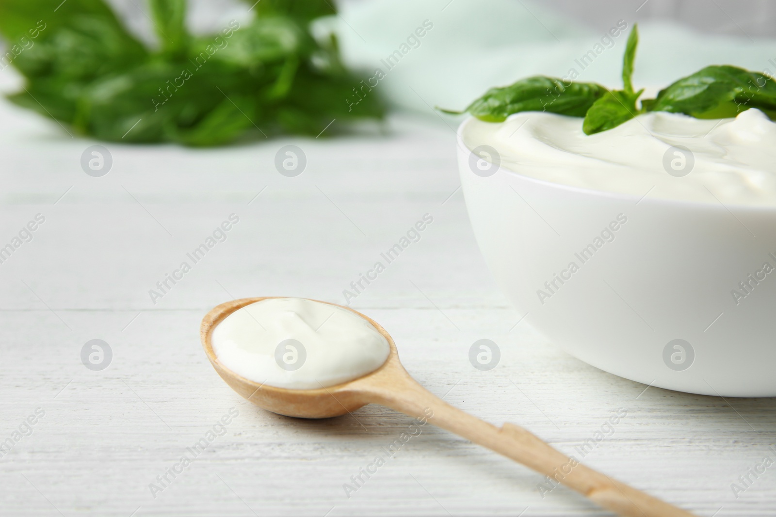 Photo of Bowl of fresh sour cream with basil and spoon on white wooden table