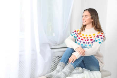 Photo of Beautiful teenage girl in warm cozy sweater looking out window at home. Space for text