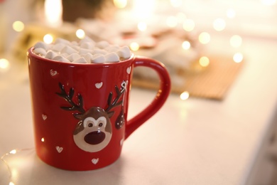 Photo of Hot drink with marshmallow and festive lights on table indoors, closeup