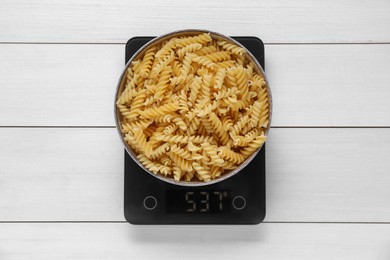 Electronic scales with uncooked pasta on white wooden table, top view