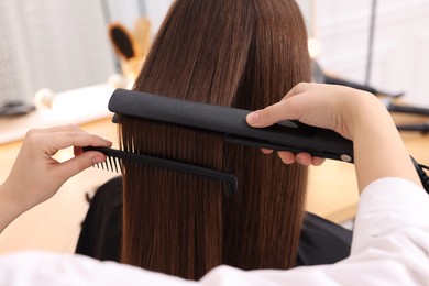 Photo of Hairdresser straightening woman's hair with flat iron in salon, closeup