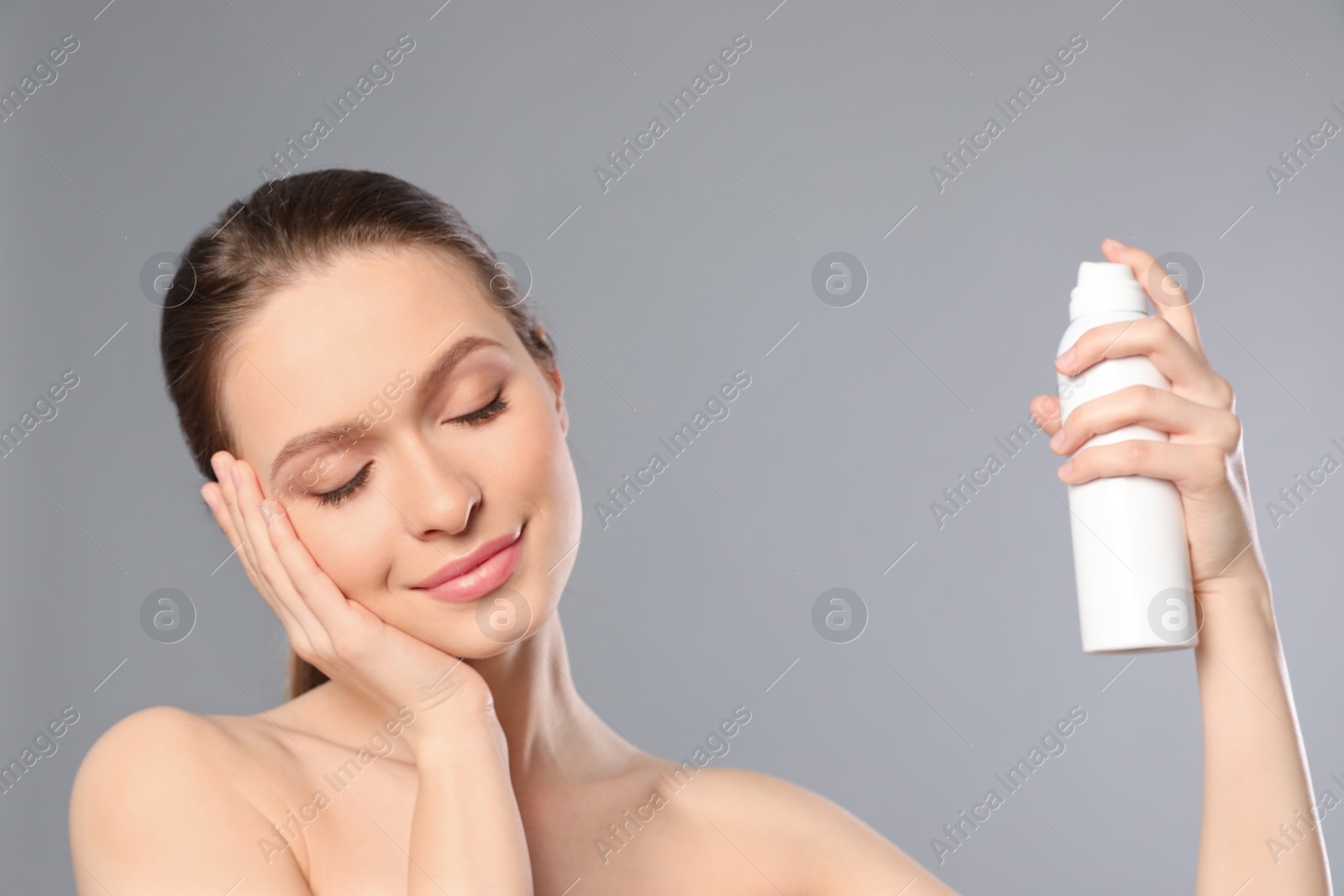 Photo of Young woman applying thermal water on face against grey background. Cosmetic product