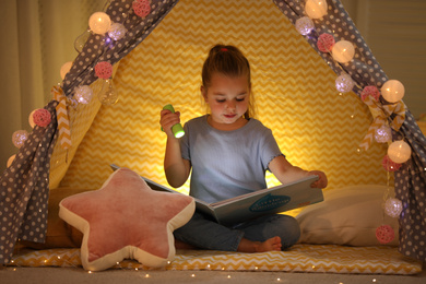 Photo of Little girl with flashlight reading book in play tent at home