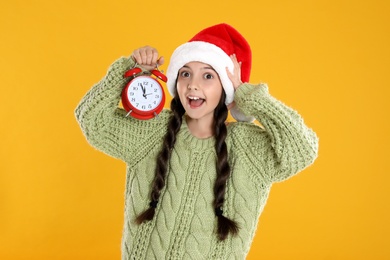 Photo of Girl in Santa hat with alarm clock on yellow background. New Year countdown