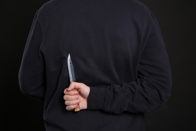 Man with knife behind his back on black background, closeup. Dangerous criminal