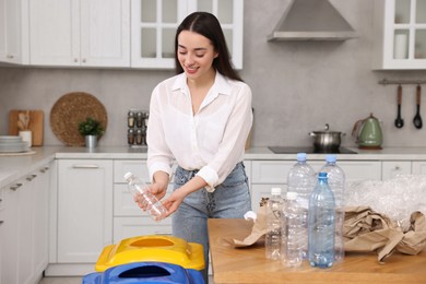 Photo of Garbage sorting. Smiling woman throwing plastic bottle into trash bin in kitchen