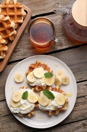 Photo of Delicious Belgian waffles with banana and whipped cream served on wooden table, flat lay