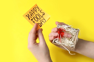 Photo of Woman holding paper with words Happy Fool's Day and fake cut finger in box on yellow background, closeup