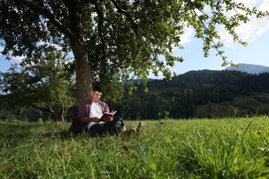 Photo of Handsome man reading book under tree on meadow in mountains