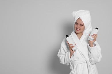 Photo of Beautiful young woman in bathrobe holding bottles of shampoo on light grey background. Space for text