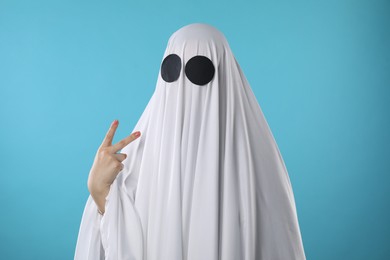Photo of Funny ghost. Woman covered with white sheet showing V-sign on light blue background