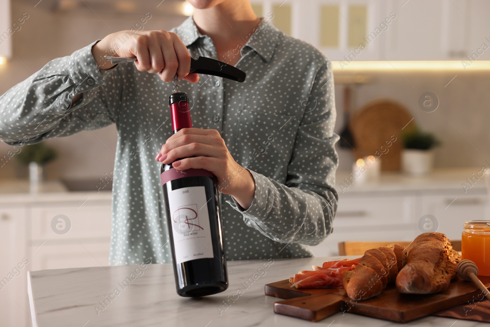 Photo of Romantic dinner. Woman opening wine bottle with corkscrew at table in kitchen, closeup