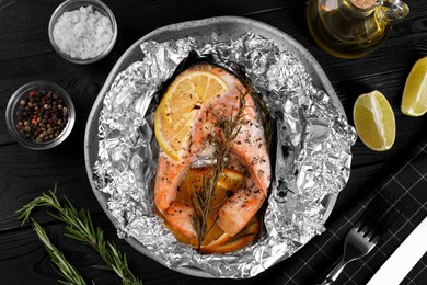 Photo of Tasty salmon baked in foil with lemon, spices and rosemary served on dark wooden table, flat lay