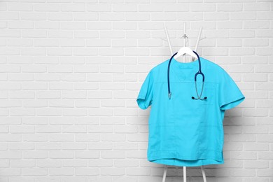 Photo of Turquoise medical uniform and stethoscope hanging on rack near white brick wall. Space for text