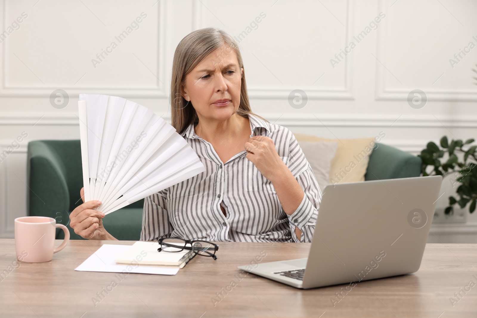 Photo of Menopause. Woman waving hand fan to cool herself during hot flash at wooden table indoors