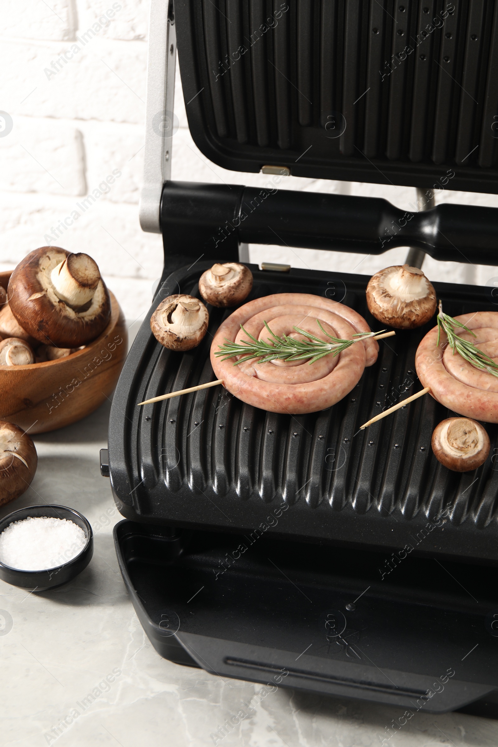 Photo of Electric grill with homemade sausages, rosemary and mushrooms on marble table