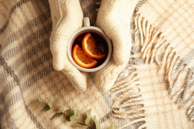 Woman with knitted mittens holding cup of hot winter drink on warm plaid, top view. Cozy season