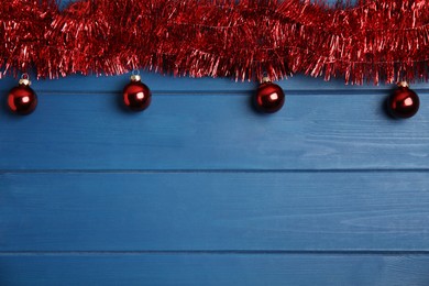 Photo of Red tinsel and Christmas balls on blue wooden background, flat lay. Space for text