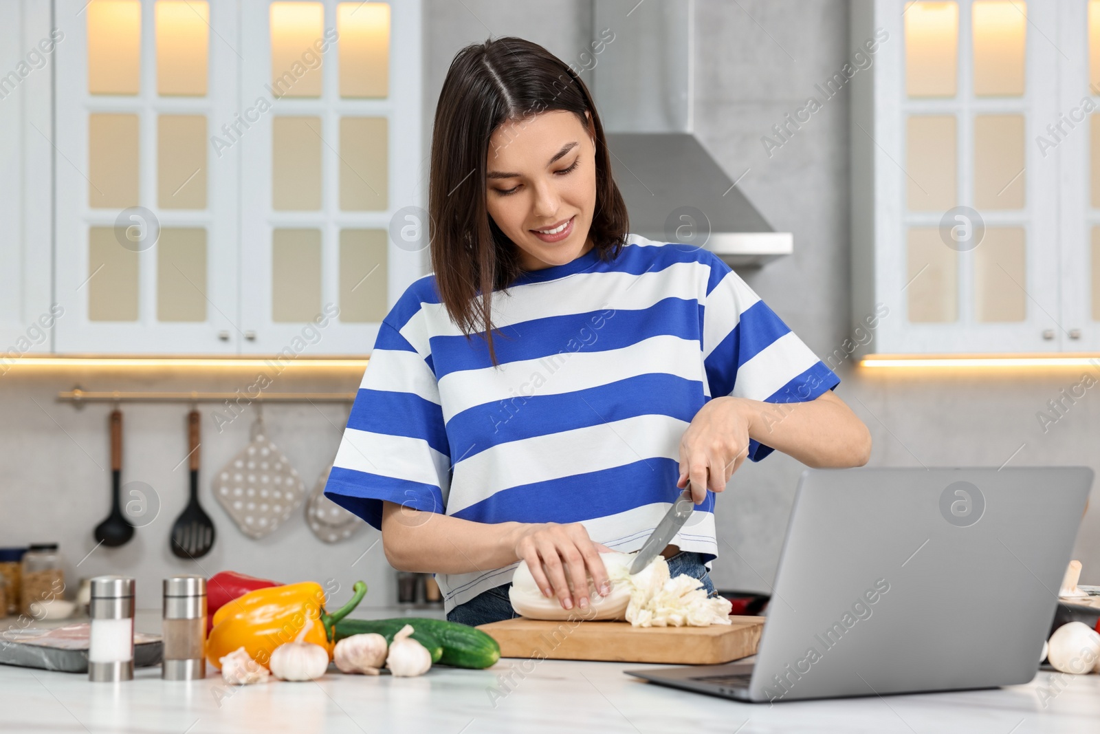 Photo of Young woman cooking while watching online course via laptop in kitchen