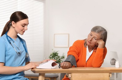 Young healthcare worker measuring senior woman's blood pressure at wooden table indoors