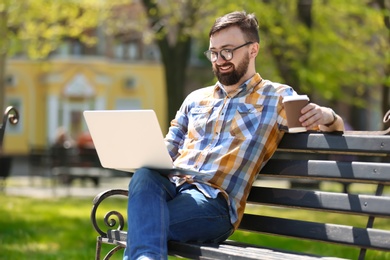 Portrait of young man with laptop and cup of coffee on bench in park