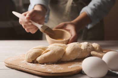 Photo of Woman spreading egg yolk onto raw braided bread at white wooden table, closeup. Traditional Shabbat challah