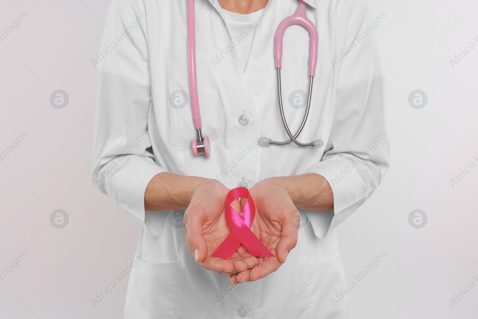 Photo of Doctor with stethoscope holding pink ribbon on white background, closeup. Breast cancer awareness