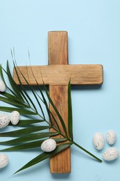 Photo of Wooden cross, painted Easter eggs and palm leaf on light blue background, flat lay