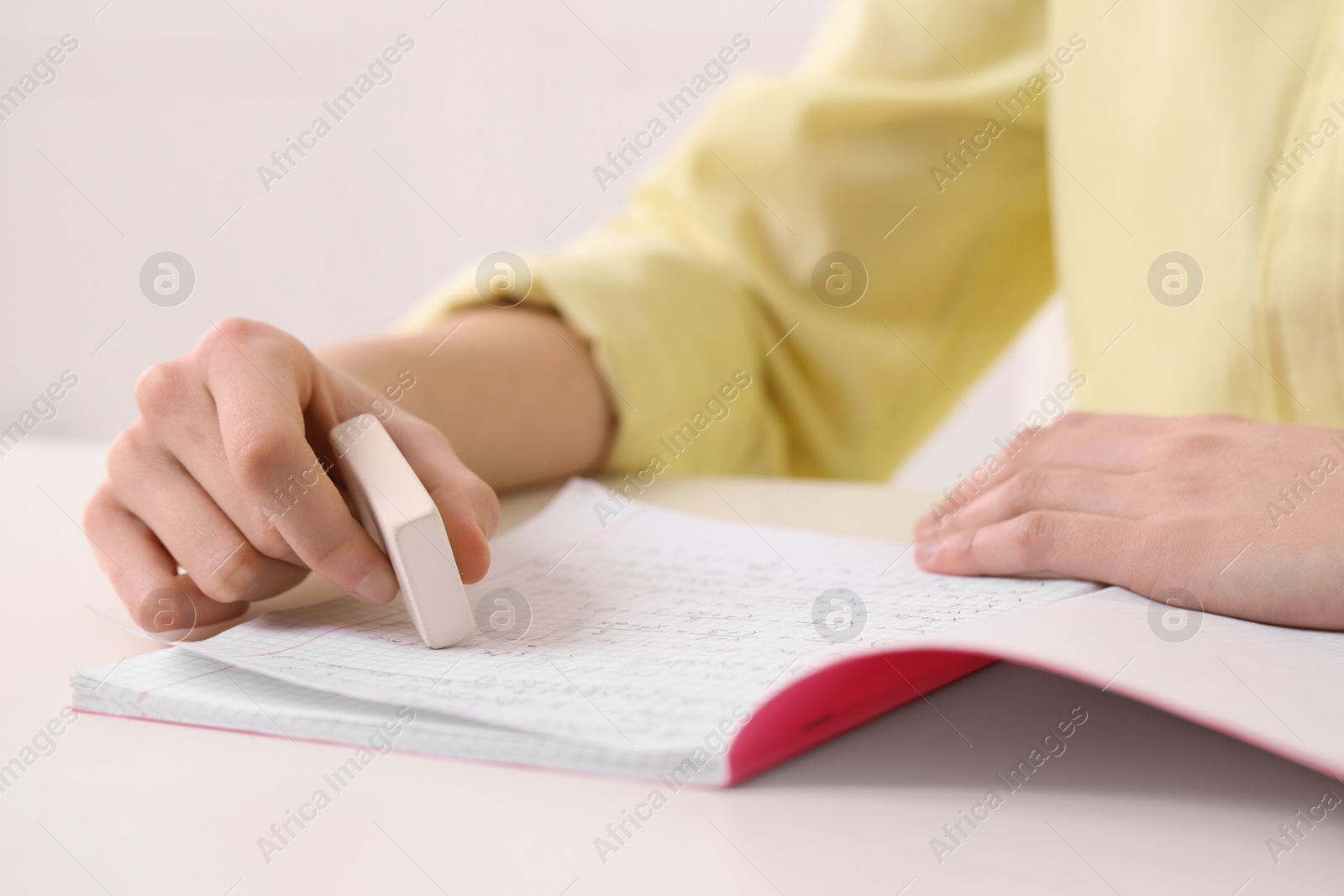 Photo of Girl erasing mistake in her notebook at white desk indoors, closeup