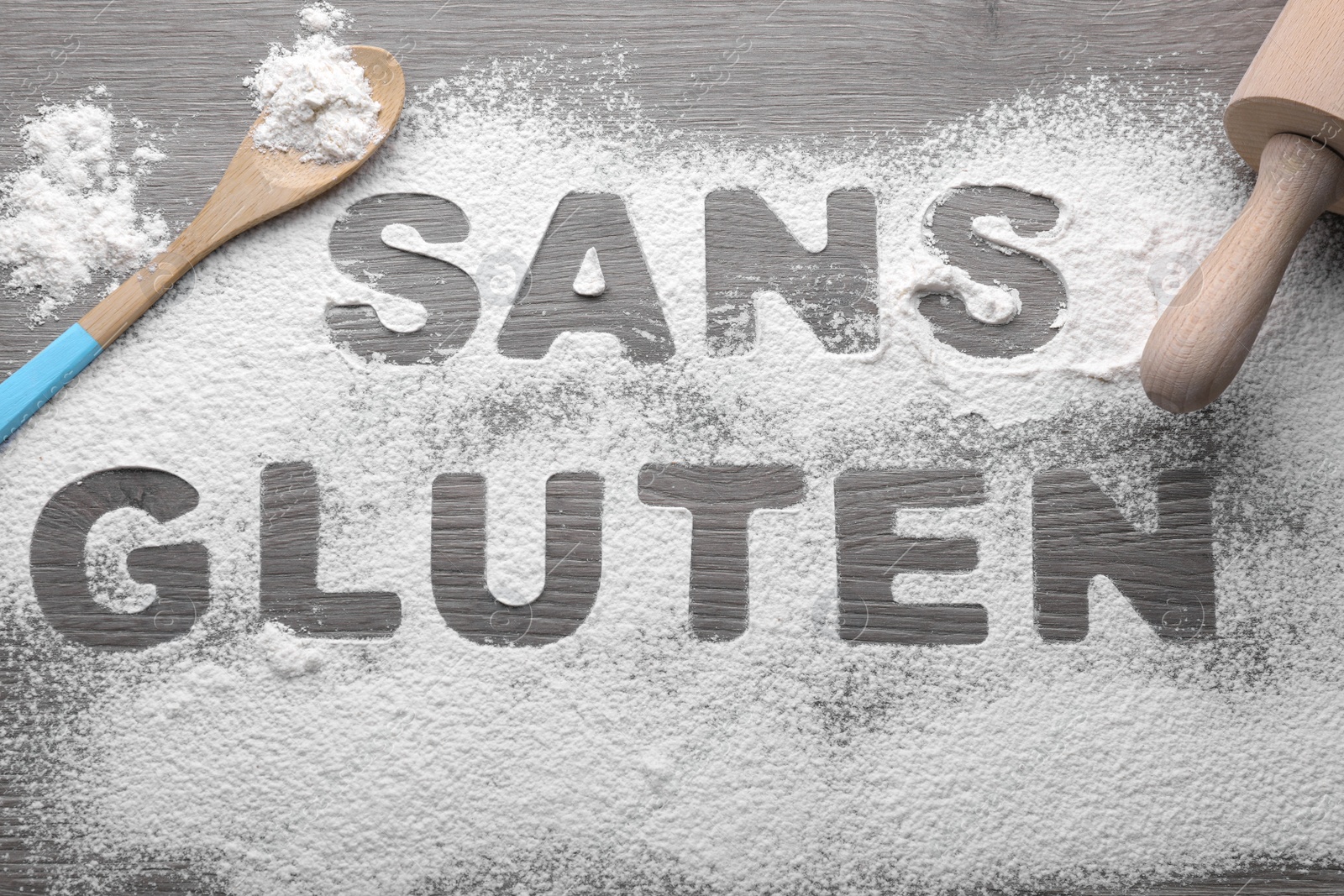 Photo of Words Sans gluten written with flour and different kitchen utensils on wooden table, flat lay