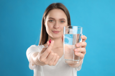 Young woman with vitamin pill and glass of water against blue background, focus on hands