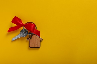 Key with trinket in shape of house and red bow on yellow background, top view. Space for text. Housewarming party