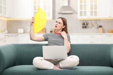 Photo of Woman with laptop waving yellow hand fan to cool herself on sofa at home