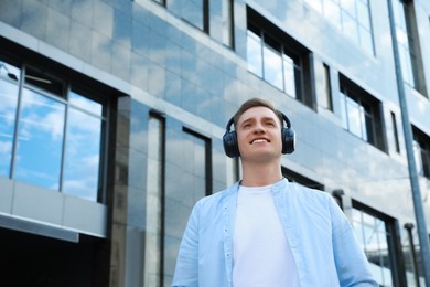 Smiling man wearing headphones near building outdoors, low angle view. Space for text