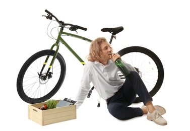 Upset young man with bottle of wine and box of belongings near bicycle on white background