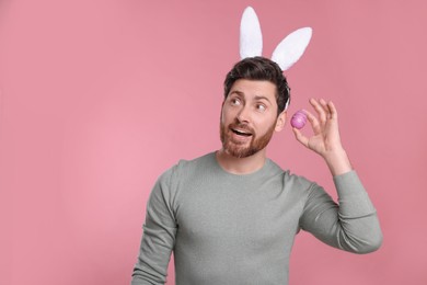 Photo of Emotional man in cute bunny ears headband holding Easter egg on pink background. Space for text
