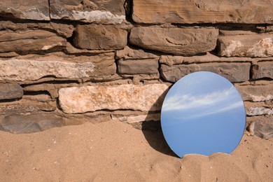 Photo of Round mirror reflecting sky on sand near stone wall outdoors, space for text