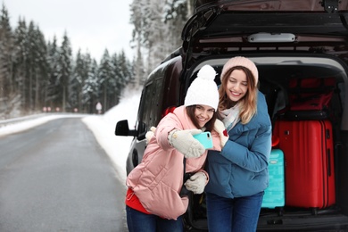 Friends taking selfie near open car trunk full of luggage on road, space for text. Winter vacation