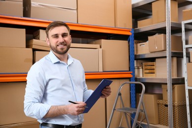 Photo of Young businessman with clipboard near rack of cardboard boxes at warehouse