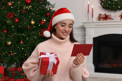 Happy young woman in Santa hat with Christmas gift box reading greeting card at home