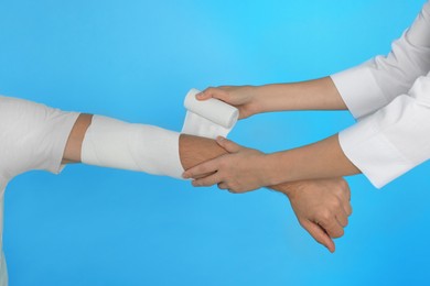 Photo of Doctor applying medical bandage onto patient's arm on light blue background, closeup