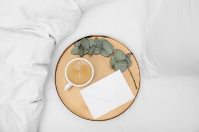 Tray with cup of coffee, envelope and eucalyptus branch on white bed, top view
