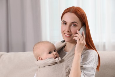 Photo of Mother talking on smartphone while holding her child in sling (baby carrier) at home