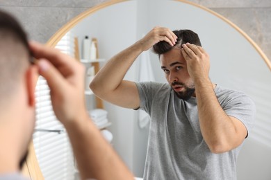 Emotional man examining his head near mirror in bathroom. Dandruff problem