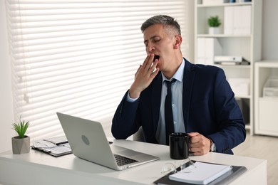 Photo of Sleepy man with cup of drink yawning at table in office