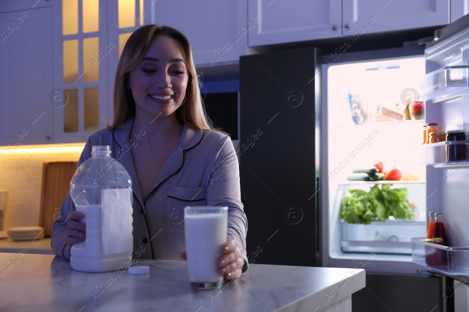 Photo of Young woman holding glass and gallon bottle of milk on white marble table in kitchen at night. Space for text