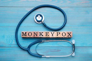Photo of Word Monkeypox made of cubes and stethoscope on light blue wooden table, flat lay