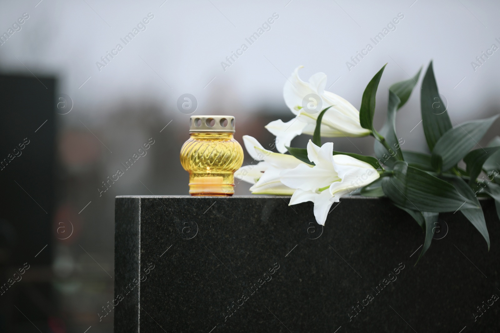 Photo of White lilies and candle on black granite tombstone outdoors. Funeral ceremony