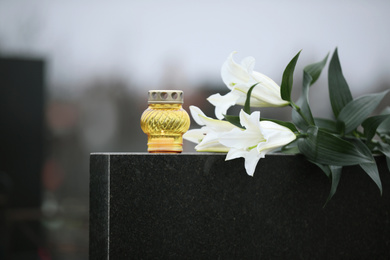 Photo of White lilies and candle on black granite tombstone outdoors. Funeral ceremony